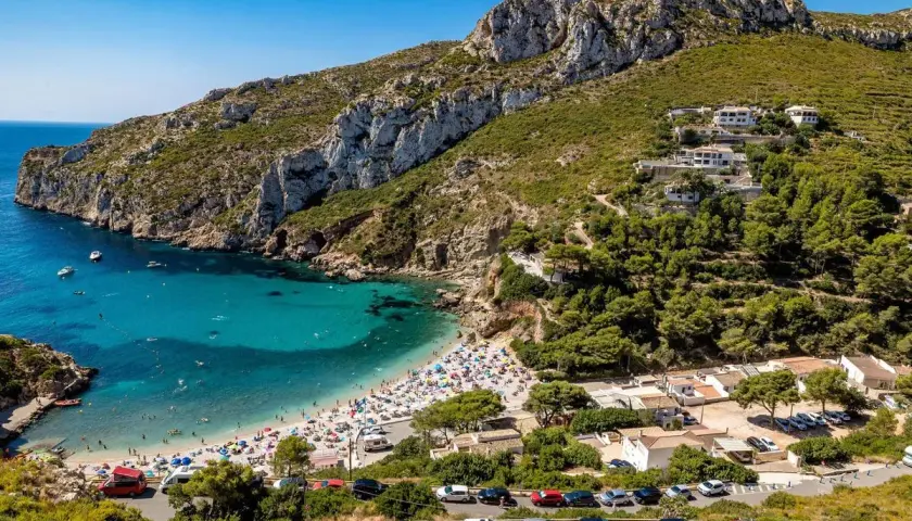 Bahía con aguas turquesas y playa abarrotada, rodeada de acantilados y vegetación. Un paraíso entre las mejores playas de Alicante.