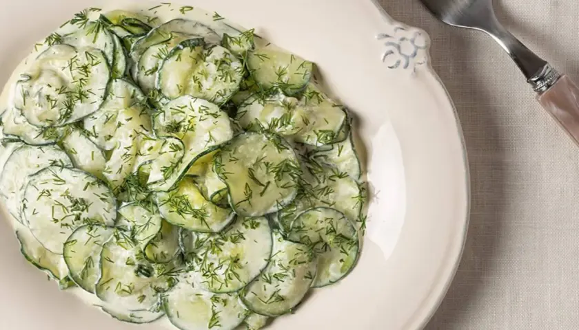 Ensalada alemana de pepino en rodajas finas, cubierta con una cremosa salsa y eneldo fresco, servida en un plato blanco.