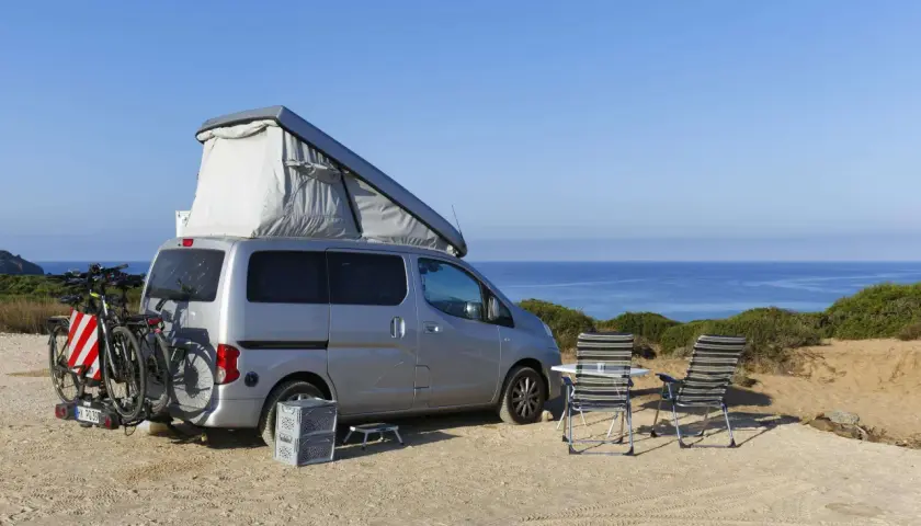 Silberner Camper mit Dachzelt und Fahrrädern am Heck, zwei Liegestühle und ein Tisch stehen bereit für entspanntes italien wildcampen am Meer.
