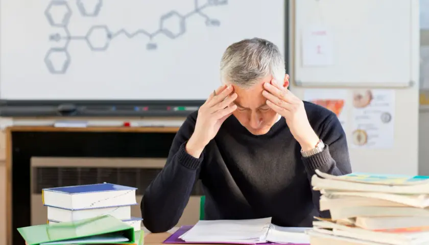 Lehrer mit Händen an den Schläfen, umgeben von Büchern und einer chemischen Formel an der Tafel. Der Lehrer-Stress ist deutlich sichtbar.