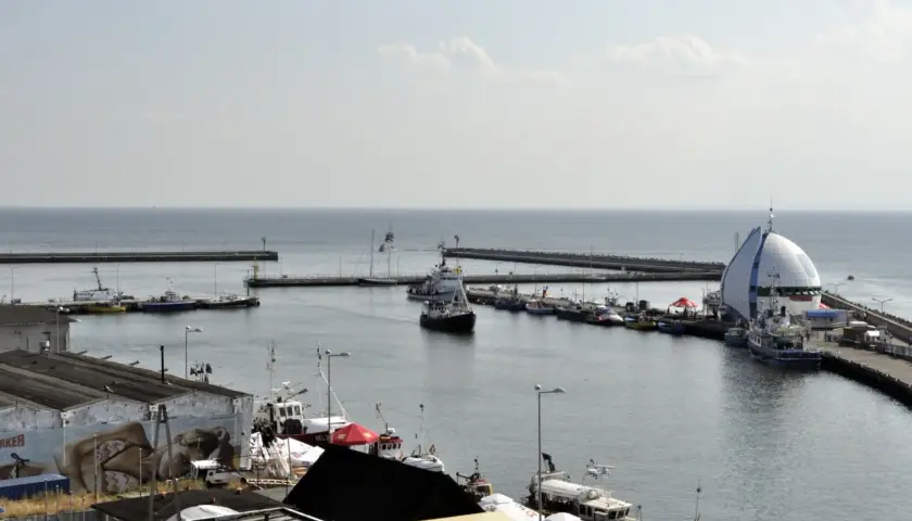 Widok na port Hel z licznymi statkami i łodziami zacumowanymi przy nabrzeżu. W tle widać morze i falochrony.