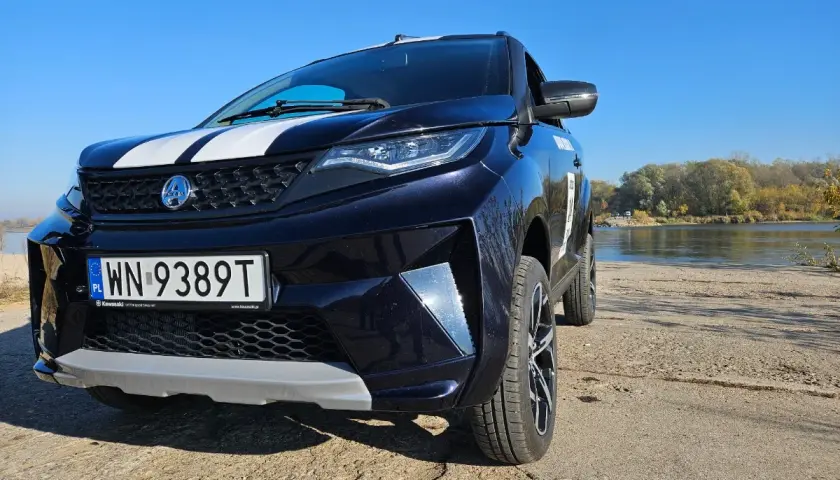 Ciemnoniebieskie aixam auto z białymi pasami na masce stoi nad rzeką w słoneczny dzień.