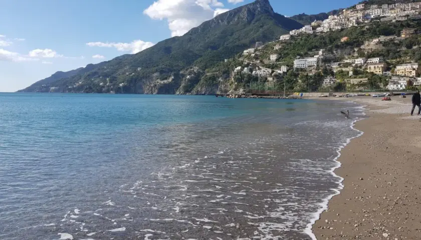 La plage de Vietri sul Mare s'étend sous un ciel bleu, avec des maisons blanches perchées sur la colline verdoyante. L'eau turquoise caresse le sable.