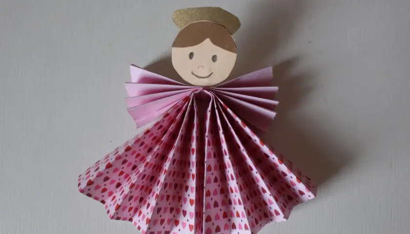 Un adorabile angelo di carta da ritagliare, con un vestito a fantasia di cuoricini rosa e un'aureola dorata.