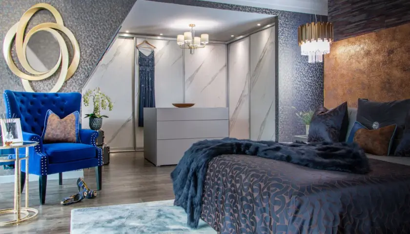 Luxurious bedroom with a king bed styled with dark blue bedding and a faux fur throw. A plush blue armchair and gold accents complete the opulent look.