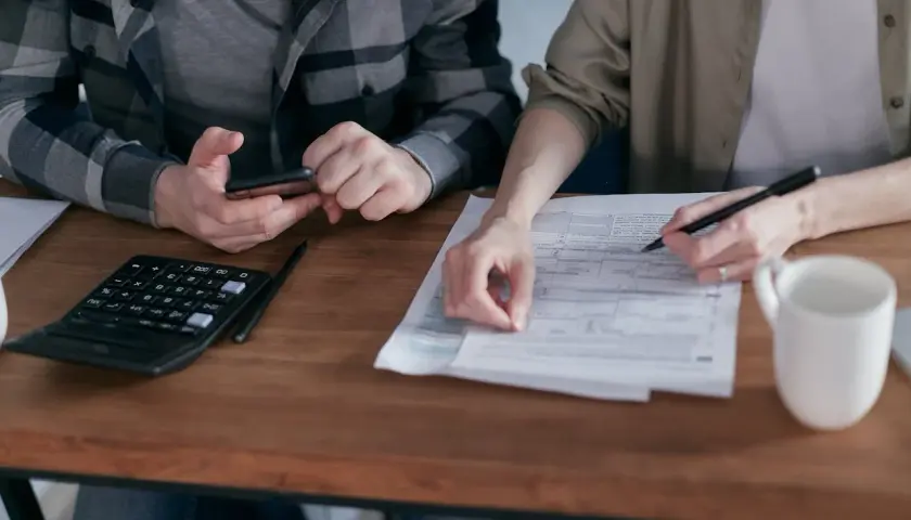 Dwie osoby przy drewnianym stole wypełniają dokumenty, obok kalkulator i kubek.