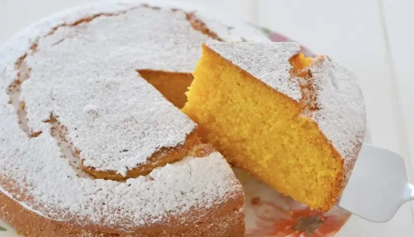Una fetta di torta di carote e yogurt, soffice e dorata, viene sollevata da una spatola. La torta è cosparsa di zucchero a velo.