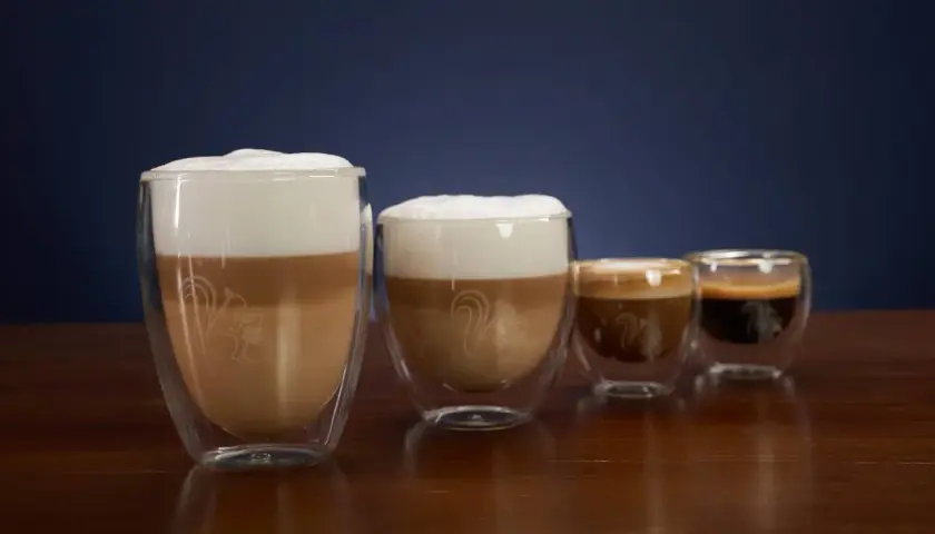Vier Gläser mit Kaffee und Milchschaum. Das größte Glas zeigt, was ist milchkaffee: geschichtete Milch und Kaffee mit viel Schaum.