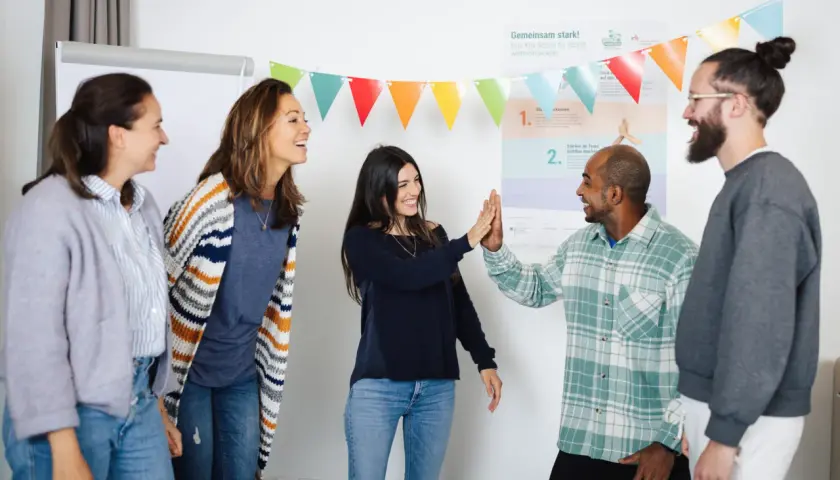 Ein motiviertes Lehrerteam feiert einen Erfolg mit High-Fives. Bunte Wimpelketten und eine Wandtafel deuten auf eine positive Lernumgebung hin.