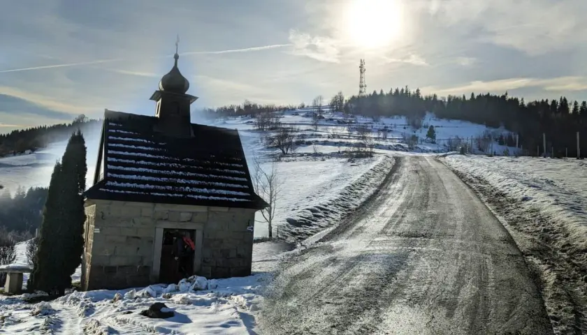 Kamienna kapliczka na zboczu ośnieżonej koskowa góra, z drogą wijącą się w słońcu.