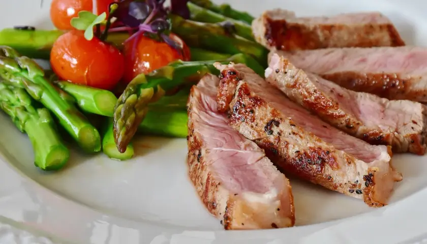 Tranches de secreto de porc grillé, accompagnées d'asperges vertes et de tomates cerises sur une assiette blanche.