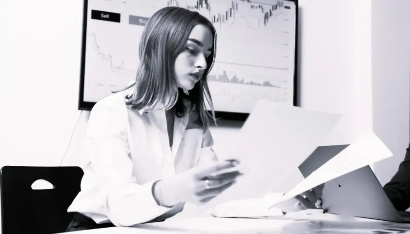Kobieta analizuje dokumenty, podczas gdy na ekranie w tle widać wykresy finansowe. Bank sprawdza co po udzieleniu kredytu, by upewnić się co do stabilności finansowej.