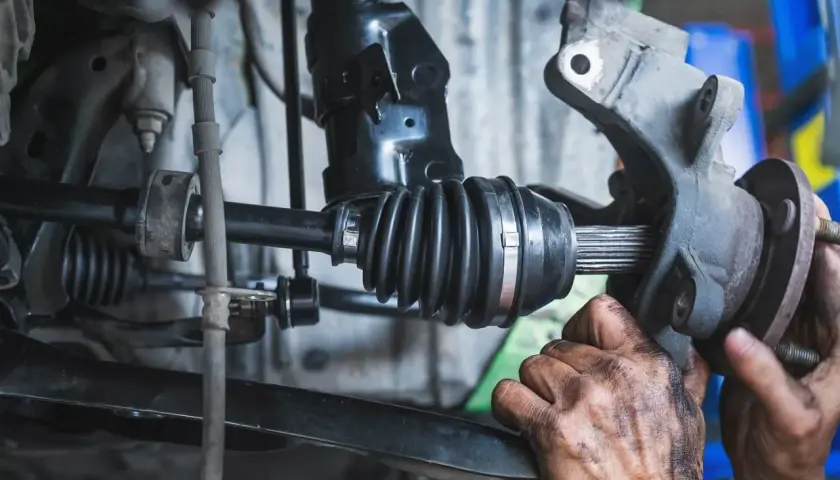 Mechanik naprawia przegub napędowy samochodu, widoczne są jego zabrudzone ręce i elementy zawieszenia.
