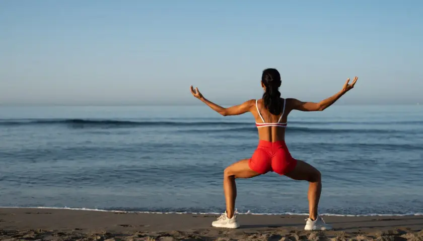 Kobieta w czerwonych szortach i sportowym topie ćwiczy na plaży. Jej szeroko rozstawione nogi i wyciągnięte ręce sugerują rozciąganie, być może angażujące powięź szeroką.
