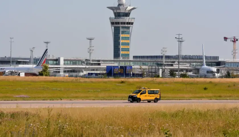 Żółty van z napisem "INSPECTION" na tle lotniska z wieżą kontroli lotów i samolotami.