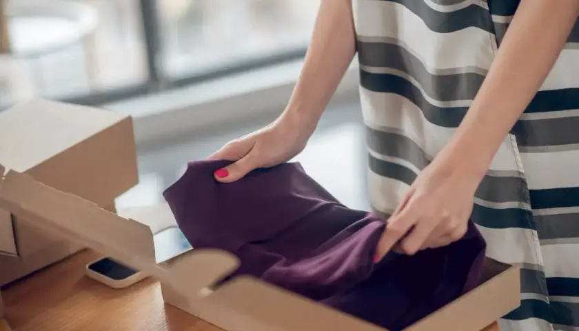 Kupujesz na Vinted i zastanawiasz się, co jeśli sprzedający nie wyśle paczki? Kobieta pakuje bordową sukienkę do kartonu.