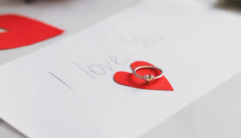Pierścionek z serduszkiem na czerwonym sercu, obok napis "I love you". Idealne życzenia walentynkowe po angielsku.