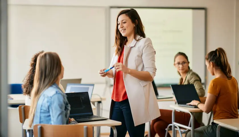Nauczycielka wyjaśnia studentom, jak zostać nauczycielem zawodu. Uczniowie pracują na laptopach w sali lekcyjnej.