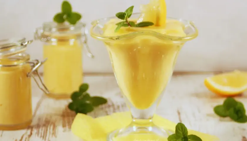Pyszny, kremowy lemon curd w pucharku, ozdobiony plasterkiem cytryny i miętą. Obok dwa słoiczki z tym samym musem.