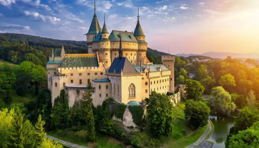 Bajkowy pałac romantyczny otoczony zielenią, z wieżami i fosą, w blasku zachodzącego słońca.