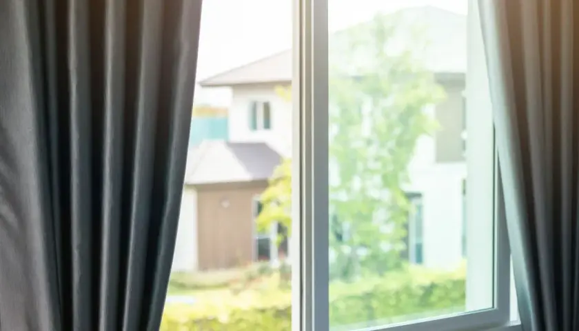 Tende grigie aperte su una finestra, lasciando intravedere case e alberi verdi. Si percepisce una leggera corrente d'aria in casa.