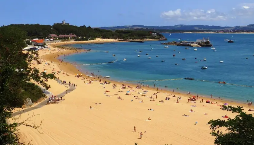La **playa de los peligros** se llena de gente disfrutando del sol y el mar azul, con barcos anclados y edificios en la costa.