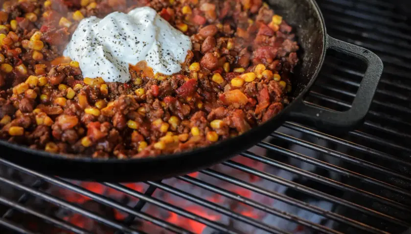 Chili con carne fumant dans une poêle sur un barbecue, garni de crème fraîche.