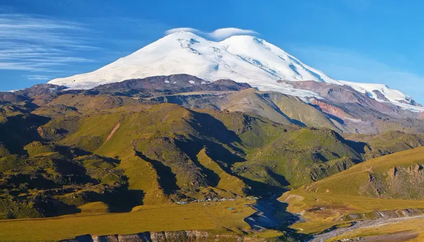 Elbrus szczyt w chmurach, otoczony zielonymi zboczami i obozem u podnóża.