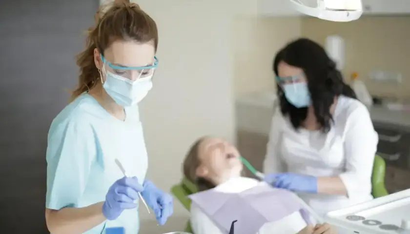 Dwie dentystki w maskach i okularach pracują przy pacjencie. Jedna z nich trzyma narzędzie, być może do usuwania pozostawionych szwów nierozpuszczalnych.