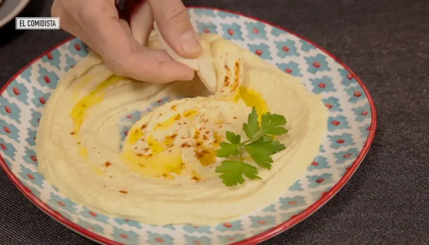 Un trozo de pan pita se sumerge en un cremoso hummus, adornado con aceite de oliva y perejil. Descubre qué es el tahini y cómo prepararlo.
