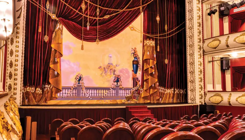 Scena teatralna z czerwonymi zasłonami, malowidłem i rzędami drewnianych krzeseł.