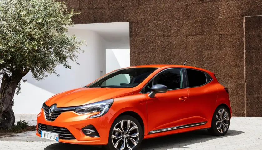 En orange Renault Clio 2026 parkerad vid en modern byggnad med en olivträd i förgrunden.