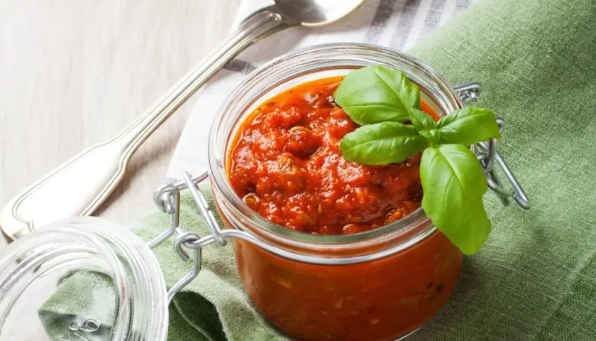 Tarro de cristal con salsa napolitana casera, decorado con hojas de albahaca fresca. Cuchara plateada y servilleta verde al lado.