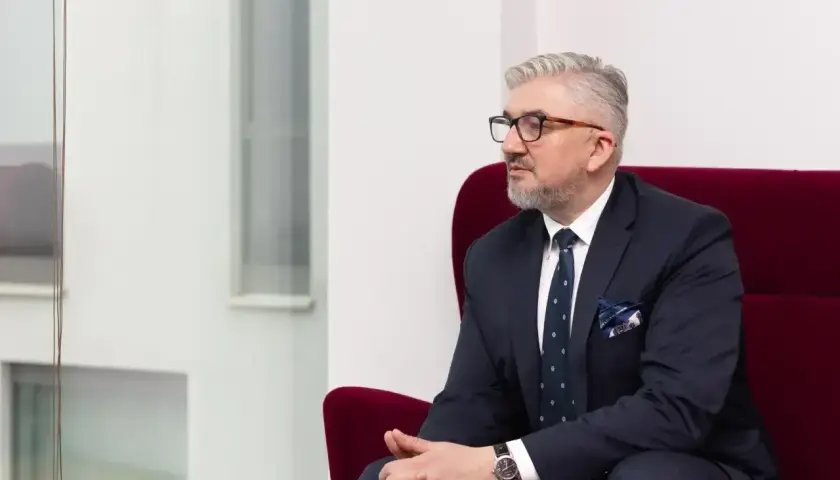 Mężczyzna w garniturze siedzi na czerwonym fotelu, patrząc w bok.