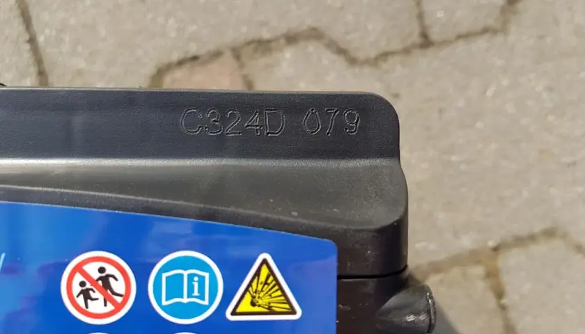Na akumulatorze Exide widoczny kod C324D 079, który może oznaczać datę produkcji. Obok symbole ostrzegawcze.