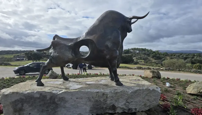 Escultura de toro en la plaza toros Valdemorillo. Paisaje rural con colinas verdes y cielo nublado.
