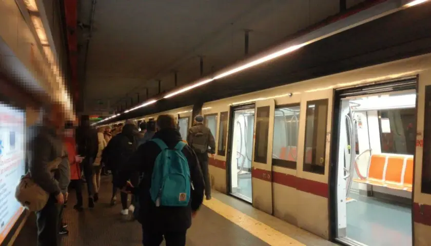 Persone in attesa sul binario della metro domenica, con un treno bianco e rosso fermo con le porte aperte.