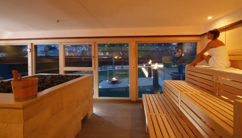 Entspannter **Sauna Bereich** mit Holzbänken und Blick auf Feuerstellen im Garten. Eine Frau in einem Handtuch sitzt auf der obersten Bank.