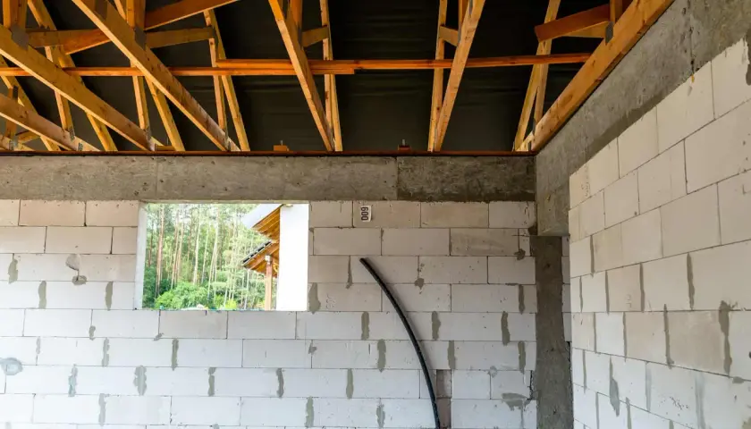 Budowa domu w toku. Widoczny wieniec stropowy, ściany z bloczków, drewniana więźba dachowa i widok na las.