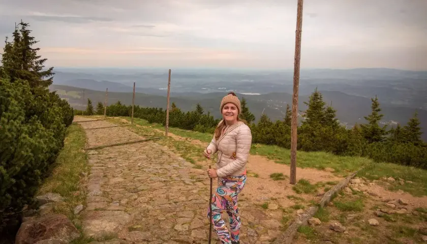 Karpacz-Śnieżka czas wejścia: uśmiechnięta turystka na kamiennym szlaku, w tle rozległa panorama gór.