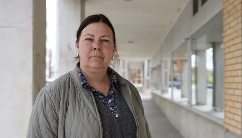 Jessica de Geer står utanför en byggnad med tegel och betong. Hon bär en grå jacka över en mönstrad skjorta och en stickad väst.