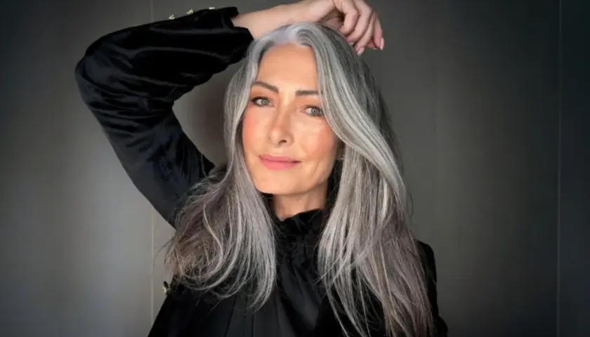 Mujer con cabello largo y ondulado, mostrando una hermosa integración de canas. Su mano acaricia su cabeza, resaltando la textura y el color natural.