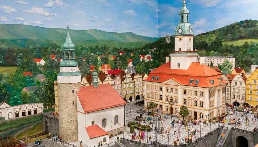 Miniaturowa panorama miasta z wieżami, ratuszem i placem pełnym postaci. W tle wzgórza i lasy, jakby z krainy jagniątków.
