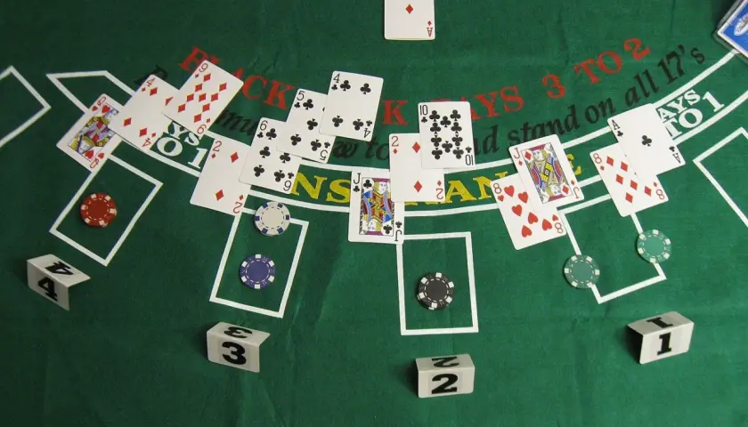 Na zielonym stole do blackjacka leżą karty, żetony i kostki. Widać karty z **kiery**, damy, walety i asy.