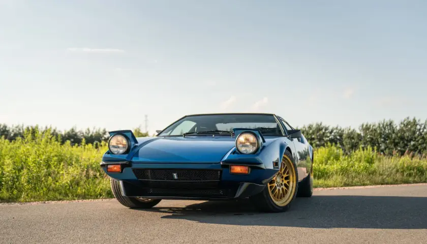 Niebieski De Tomaso Pantera z wysuwanymi światłami i złotymi felgami na tle zieleni i błękitnego nieba.