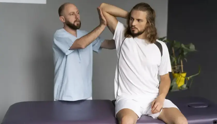 Fizjoterapeuta bada naciągnięty bark młodego sportowca siedzącego na kozetce.