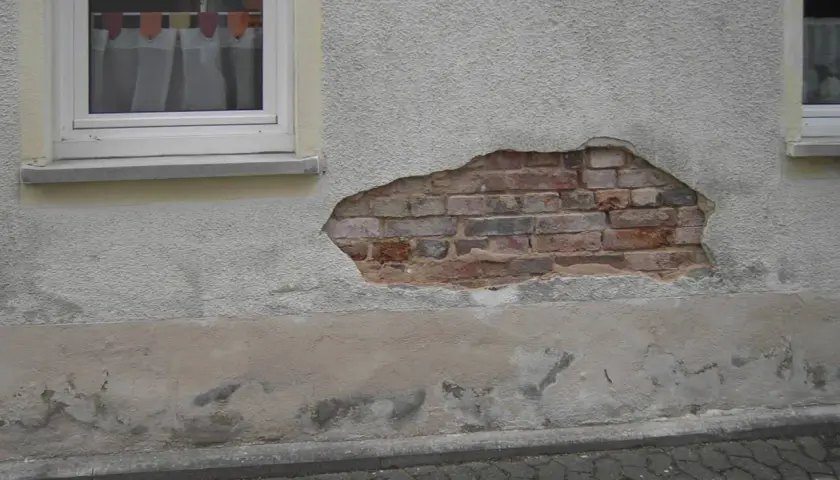 Putz löst sich im Sockelbereich, darunter Ziegelmauerwerk. Anzeichen für aufsteigende Feuchtigkeit im Sockelbereich.