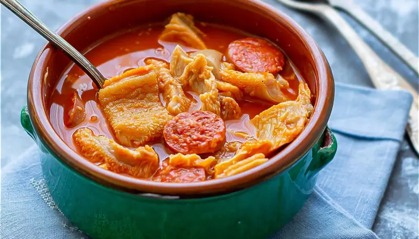 Un tazón de barro con callos a la madrileña, trozos de chorizo y un caldo rojizo. Una cuchara metálica está dentro.