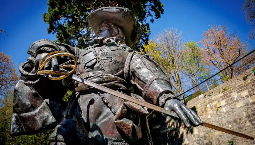 Bronzestatue eines Musketiers bei Dumas, mit Schwert und Hut, vor blauem Himmel und Bäumen.