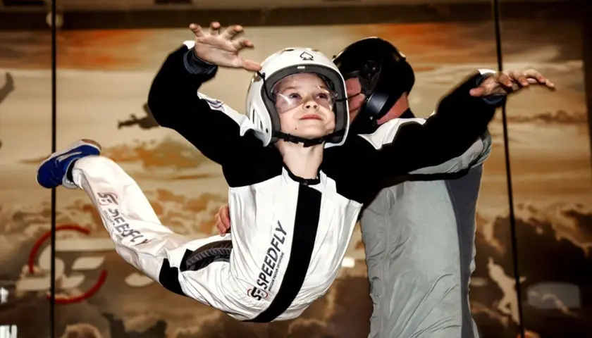Chłopiec w kasku i kombinezonie doświadcza lotów w tunelu aerodynamicznym, unosząc się w powietrzu pod okiem instruktora.